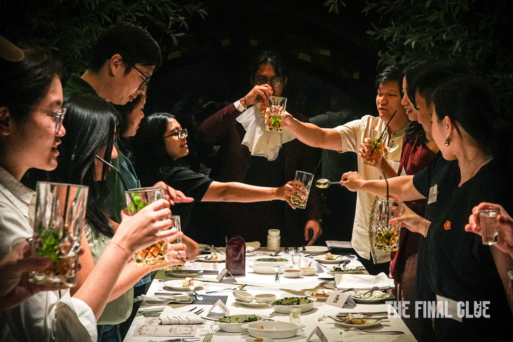 A long dinner table mid-service at a Final Clue event, guests in character across plates, glassware and printed clue cards.